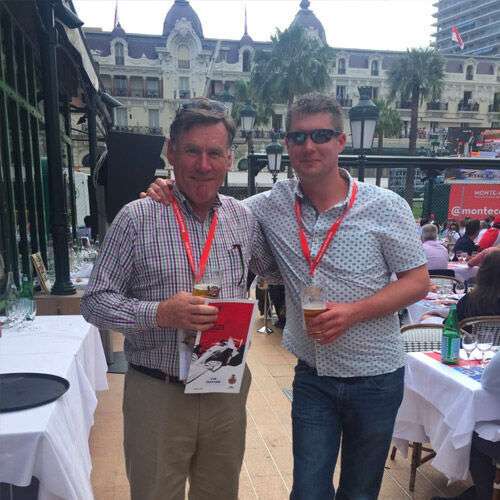 Angus MacKinnon (left) and Stuart MacKinnon (right) smiling at the camera holding drinks. Stuart has his arm around Angus's shoulders.